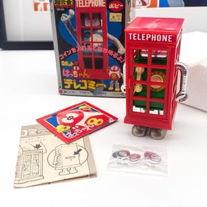 Puede incluir: Un teléfono de juguete rojo con un cuerpo de robot y una ranura para monedas. El juguete está etiquetado como "TELEPHONE" y tiene una pequeña figura roja con el número "8" en él. El juguete viene con instrucciones y una bolsa de monedas.