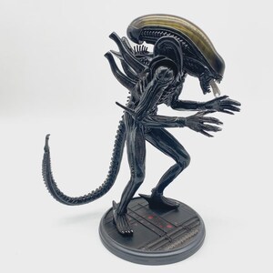 May include: A black Xenomorph figurine standing on a circular base with a textured surface. The creature features an elongated translucent head, a long segmented tail, and sharp claws. The figure is posed in a crouched position with its mouth open.