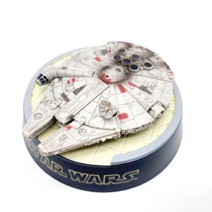 Può includere: Un modello di nave spaziale Millennium Falcon bianco e rosso su una base nera con la scritta "STAR WARS" in lettere dorate.