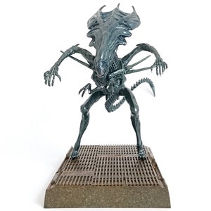 Op de afbeelding: Een gedetailleerd, donkergrijs Alien Xenomorph figuur, staand op een getextureerd platform. Het wezen heeft een lange, gesegmenteerde staart, een langwerpig hoofd en meerdere ledematen. Het figuur poseert met uitgestrekte armen, wat het dreigende ontwerp laat zien.