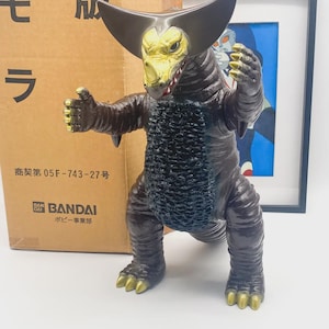Peut inclure: Figurine en vinyle Bandai marron et or représentant un monstre avec un visage jaune, des cornes noires et des griffes dorées. La figurine est dans une pose dynamique. Une boîte en carton avec du texte japonais est en arrière-plan.