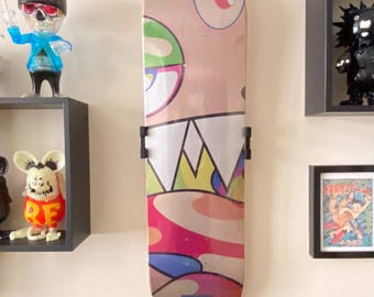 May include: A skateboard with a colorful cartoon graphic of a face with large eyes, a wide mouth, and a crown. The board is mounted on a white wall with other decorative items.