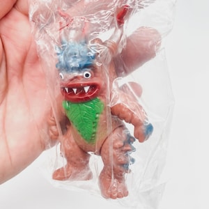 May include: A vintage monster toy figure, packaged in a clear plastic bag. The figure is primarily reddish-brown with blue accents on the head and feet. It has a green leaf-shaped chest and a red mouth and horns. The toy has a retro aesthetic.