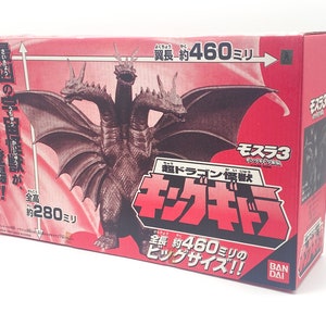 Peut inclure: Une boîte rouge avec une image d'un monstre à trois têtes avec des ailes. La boîte indique "Mothra 3" et "Ultra Dragon Beast King". Le monstre mesure environ 460 millimètres de long et 280 millimètres de haut.