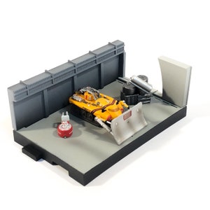 Pode incluir: Um veículo de brinquedo amarelo com uma pá, etiquetado como "FIREFLY", está estacionado em um ambiente de garagem em miniatura. A garagem é feita de papelão cinza e tem uma cerca cinza. Há vários barris pequenos cinzas e um cano cinza no fundo.