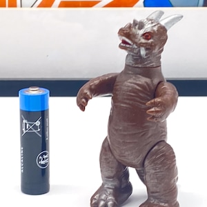 Peut inclure: Un monstre en jouet brun et gris avec une corne argentée et des yeux rouges. Le monstre est debout sur une surface blanche à côté d'une pile alcaline noire.