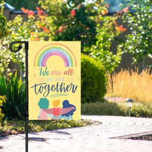May include: A yellow garden flag with a rainbow and the text "We are all in this together". Three people are holding hands in front of the rainbow.