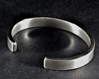 May include: A silver-toned cuff bracelet with a smooth, brushed metal finish. The bracelet features a simple, open-ended circular design with rounded edges, showcasing a clean, reflective surface that catches the light along its curved band.