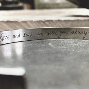 Op de afbeelding: Een zilverkleurige metalen manchetarmband met een geborstelde afwerking. Het binnenoppervlak is voorzien van de gegraveerde zwarte cursieve tekst, love and luck are with you always, gecentreerd over de lengte van de band.
