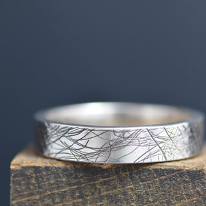 May include: A silver band with a textured, etched design. The band is sitting on a wooden surface.