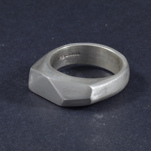 May include: A silver ring with a geometric, faceted design. The ring is made of brushed metal and has a matte finish.