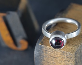 May include: A silver ring with a round, dark red gemstone set in a bezel setting.