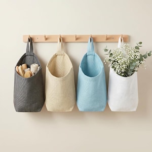 Puede incluir: Cuatro bolsas de almacenamiento colgantes de tela en gris oscuro, beige, azul claro y blanco, suspendidas de un perchero de madera clara. La bolsa gris oscuro contiene cepillos de madera, mientras que la blanca contiene follaje verde.