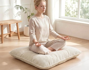 Puede incluir: Cojín de meditación de pana beige. El cojín cuadrado tiene una textura acanalada y está colocado sobre un suelo de madera clara. Una persona está sentada en el cojín en posición meditativa, con los ojos cerrados. El fondo incluye una ventana, una planta y un taburete de madera.
