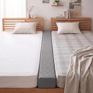 Puede incluir: Un puente de cama rectangular gris con un patrón de diamantes acolchado colocado entre dos camas individuales. La cama izquierda tiene una sábana y almohada blancas lisas, mientras que la derecha tiene una sábana a cuadros gris y blanca.