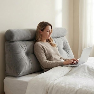 Op de afbeelding: Een grijs, gecapitonneerd bedkussen met een cilindrische hoofdsteun en een zak aan de zijkant. Het kussen ligt op een bed met een wit dekbed. Een persoon gebruikt een laptop terwijl hij tegen het kussen leunt.