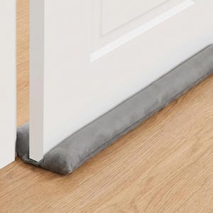 May include: A long, cylindrical, grey fabric door draft stopper positioned horizontally along the base of a white interior door. The soft, padded textile accessory rests flush against the bottom edge of the door to cover the gap between the door and the floor.