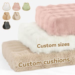 May include: Three fluffy, rectangular cushions in beige, white, and pink are stacked. The cushions have a soft, furry texture. The words "Custom sizes" and "Custom cushions" are printed on the image. Small, bunny-shaped cushions in various colours are also shown.