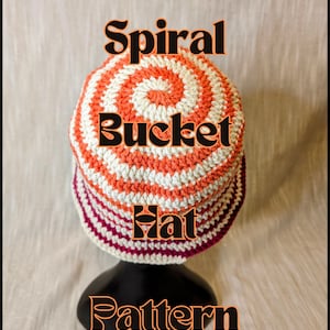 May include: A crocheted bucket hat with a spiral pattern in orange and white. The hat is on a black stand. The text "Spiral Bucket Hat Pattern" is written in black letters on a white background.