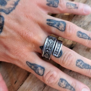 Può includere: Un anello avvolgente in metallo color argento con finitura ossidata e motivi floreali e a volute in rilievo. L'anello presenta una fascia larga che si sovrappone, mostrando intricati dettagli botanici sulla superficie del metallo.