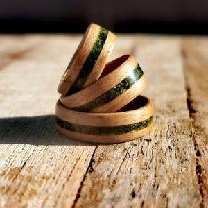May include: Three stacked rings crafted from light-toned wood, each featuring a central horizontal inlay of textured, dark green material. The rings are arranged in a vertical formation, showcasing the smooth wooden grain and the contrasting green band.