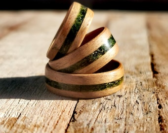 May include: Three stacked rings crafted from light-toned wood, each featuring a central inlay of textured, dark green material. The rings are arranged in a vertical formation, showcasing the smooth wooden grain and the contrasting green band.
