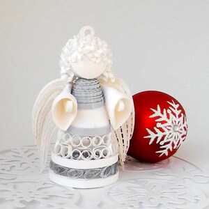 May include: White paper angel ornament with gray and white striped dress and white wings. The angel is sitting on a white snowflake-shaped base. A red Christmas ornament with a white snowflake design is in the background.