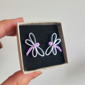 May include: A pair of white paper quilled dragonfly earrings with purple accents in a small gift box.