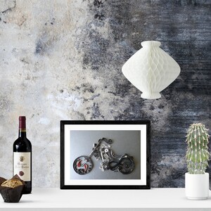 May include: A black and white framed print of a motorcycle made from paper. The motorcycle is on a grey background. The print is on a white shelf with a bottle of wine and a potted cactus.