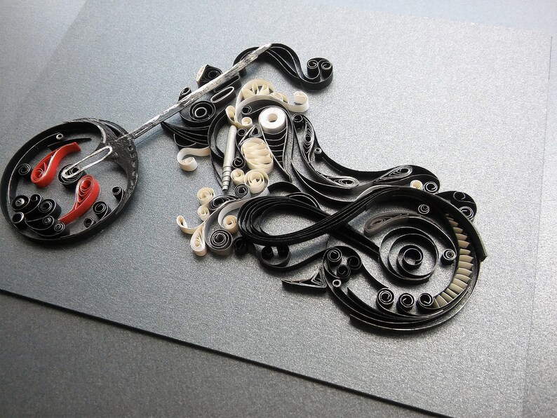 May include: A black and white paper quilling artwork depicting a motorcycle. The motorcycle is made of curled paper strips and has a red and white wheel.