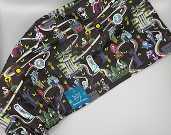 May include: A black scrub cap with a colorful, repeating pattern of cartoon ghosts, tombstones, and haunted house imagery. The cap has a blue tag with white text that reads "Cynful Creations". The cap has a yellow elastic band.