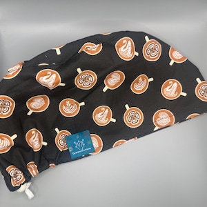 May include: Black surgical scrub cap with a repeating pattern of brown coffee cups with latte art. The latte art includes swans, hearts, and other designs. A small blue tag with white text is attached to the cap.