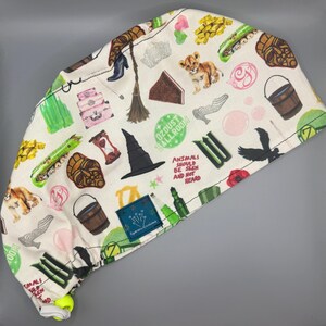 May include: White surgical scrub cap with a repeating pattern of whimsical illustrations. The design includes a lion cub, witch hat, broom, and the text "Animals Should Be Seen and Not Heard". The cap has a yellow adjustable toggle.