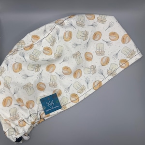 May include: White surgical scrub cap with a repeating pattern of watercolor bread, butter, and whisks. The cap has a blue label with the text "Cystoponic Creations". The cap is designed to be worn in a medical or culinary setting.