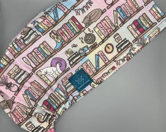 May include: A patterned scrub cap featuring a bookshelf design with books, a cat, and other decorative elements. The cap is primarily white with pink, blue, and brown accents. The design includes text such as "Midnights" and "Folklore".