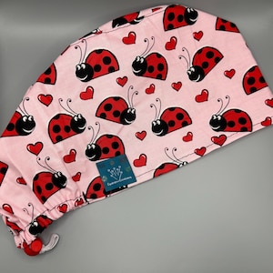 May include: A pink surgical scrub cap featuring a repeating pattern of red ladybugs with black spots and red hearts. The cap has a drawstring and a small tag with text.