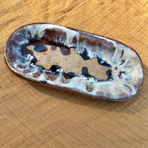 May include: An oblong ceramic dish with a speckled tan interior and a dark brown, blue, and white glazed rim. The dish has rounded edges and a glossy finish, suitable for serving or display.