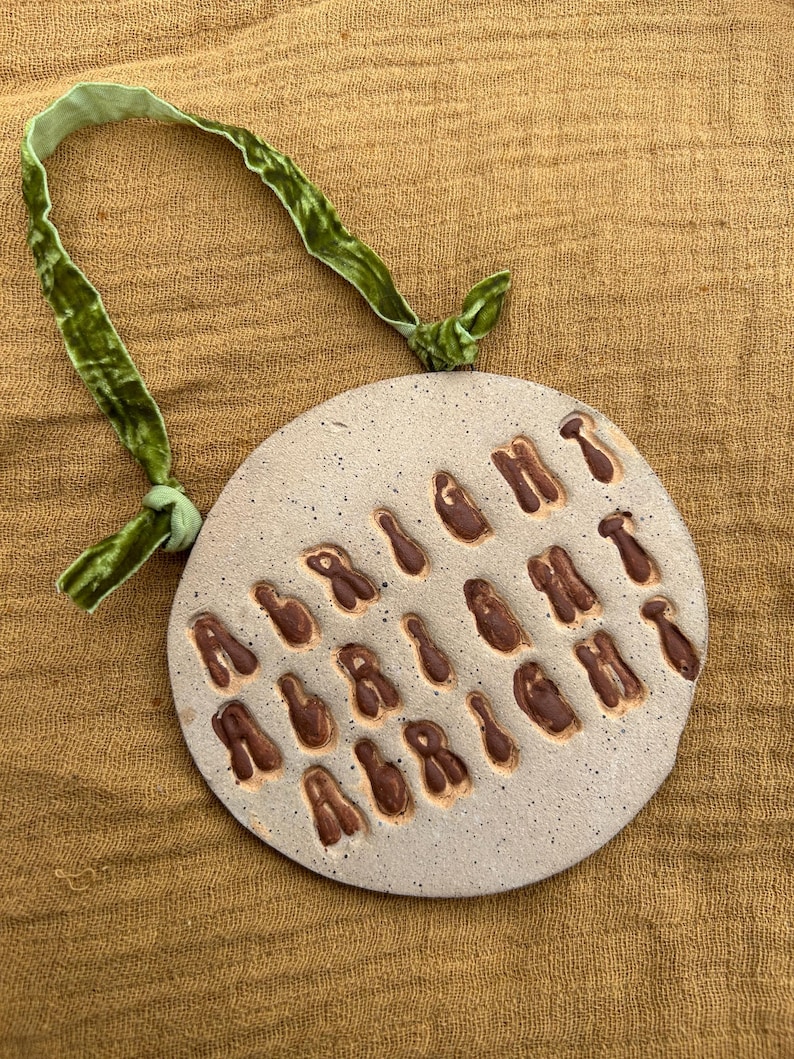 Puede incluir: Adorno redondo de cer&aacute;mica moteada con letras marrones en relieve, suspendido por una cinta de terciopelo verde. El fondo es una tela texturizada de color beige.