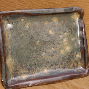 May include: A rectangular ceramic serving dish with a speckled green and brown glaze. The dish has a raised rim and a slightly uneven surface.