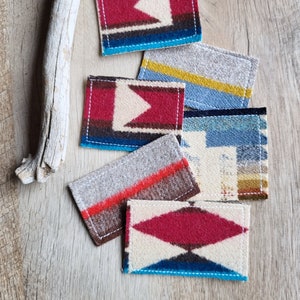 May include: A collection of rectangular coasters with colorful geometric patterns. The coasters feature designs in red, blue, beige, and gray, with white stitching around the edges. They are arranged on a wooden surface.