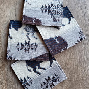 May include: A set of five square coasters with a Southwestern design. The coasters feature a repeating pattern of bison, geometric shapes, and a color palette of white, gray, and navy blue. The edges are finished with a dark brown trim.