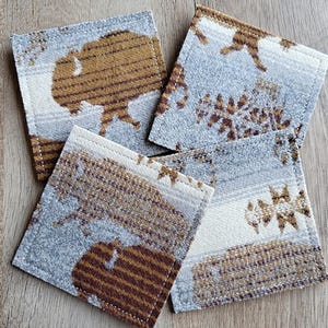 May include: Set of four square coasters with a Southwestern design. Each coaster features a repeating pattern of a bison in shades of brown and tan against a light blue and white background. The coasters are edged with white stitching.