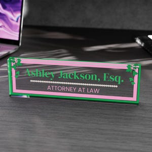 May include: A clear acrylic desk nameplate with a pink and green border. The nameplate features the name "Ashley Jackson, Esq." in green text, along with the words "ATTORNEY AT LAW". Decorative green ivy accents the corners.