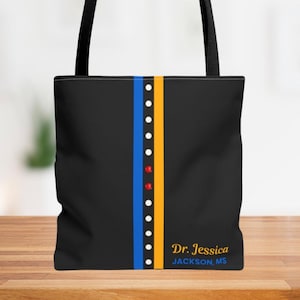 Op de afbeelding: Zwarte draagtas met verticale blauwe en gele strepen, geaccentueerd met witte en rode stippen. De tas heeft de tekst "Dr. Jessica JACKSON, MS" in gouden letters. De tas heeft zwarte handvatten.