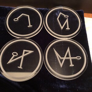 May include: Four round, clear glass coasters with white outlines and different geometric designs inside. The coasters are on a dark blue velvet surface.