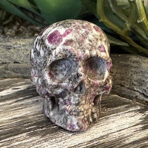 May include: A carved skull figurine made of a stone material. The skull is primarily gray with patches of white and dark pink. The eye sockets, nose, and mouth are clearly defined. The skull is a decorative item.