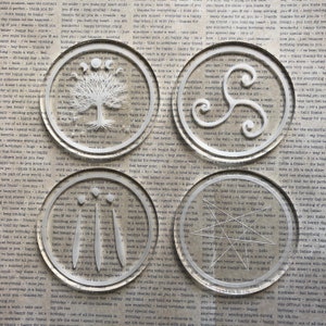 May include: Set of four silver coasters with different designs. Each coaster features a white, intricate design on a silver background. The designs include a tree with roots, a Celtic knot, a star, and a moon with three phases.