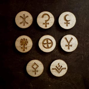 May include: Nine circular wooden tokens, each engraved with a different symbol. The symbols include a crescent moon, a cross within a circle, and other geometric designs. The tokens are light brown and are on a dark brown surface.