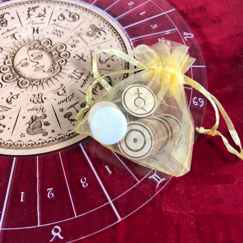 K&ouml;nnte beinhalten: Ein Holz-Zodiakrad mit einer transparenten Plastikabdeckung und einem gelben Organzas&auml;ckchen, das Holzm&auml;rkchen mit astrologischen Symbolen enth&auml;lt. Die Marken beinhalten ein Symbol f&uuml;r den Planeten Jupiter, ein Symbol f&uuml;r die Sonne und ein leeres Marken.