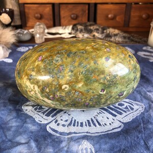 May include: A large, smooth, green and brown egg-shaped stone with pink and white flecks. The stone is sitting on a blue fabric with a white butterfly design.